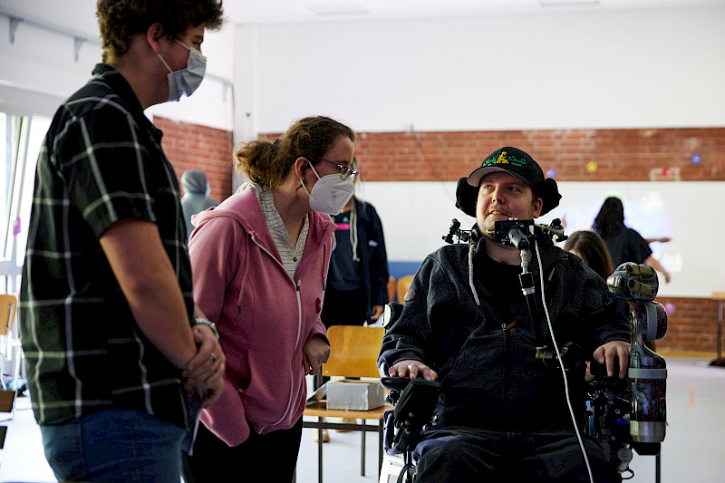 Zwei Jugendliche sprechen mit einem Erwachsenen in einem Rollstuhl. Er trägt eine Cappy mit der Aufschrift wheelyworld.