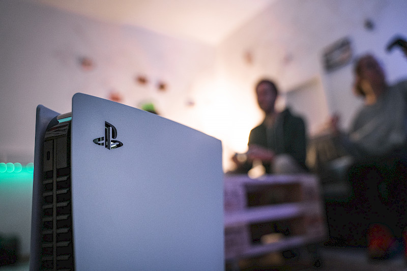 Die Playstation 5 im Fokus. Im Hintergrund sind verschwommen zwei Spieletester