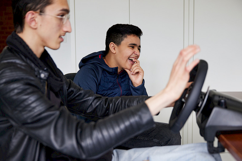 Ein Jugendlicher spielt mit einem Lenkrad. Ein anderer Jugendlicher sitzt dahinter und lacht.