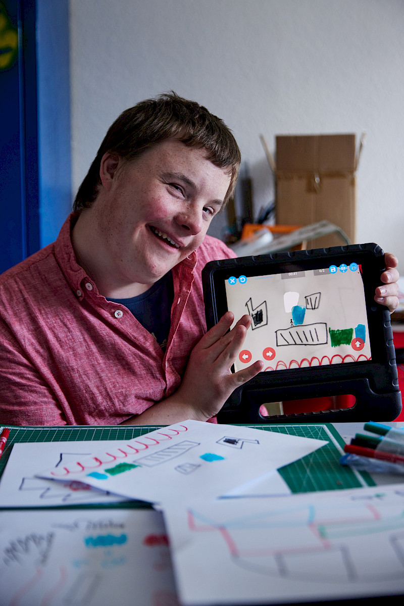 Ein Jugendlicher hat ein Jump'n'Run Spiel gemalt. Er hält ein iPad hoch. Er zeigt sein Spiel. Die App heißt Draw your Game.