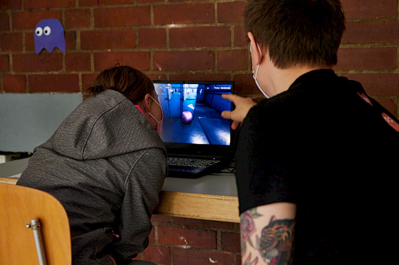 Eine Jugendliche steuert ein Spiel auf einem Laptop mit den Augen. Ein Erwachsener unterstützt sie und zeigt auf den Bildschirm.