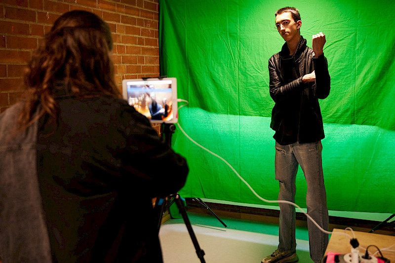 Ein Jugendlicher posiert vor einem Greenscreen. Eine Erwachsene macht davon ein Foto mit einem iPad.