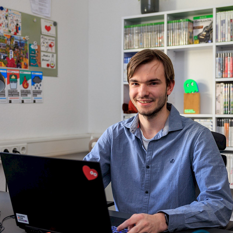 Martin sitzt vor einem Laptop. Er trägt ein blaues Hemd und hat kurze braune Haare. Er lächelt.