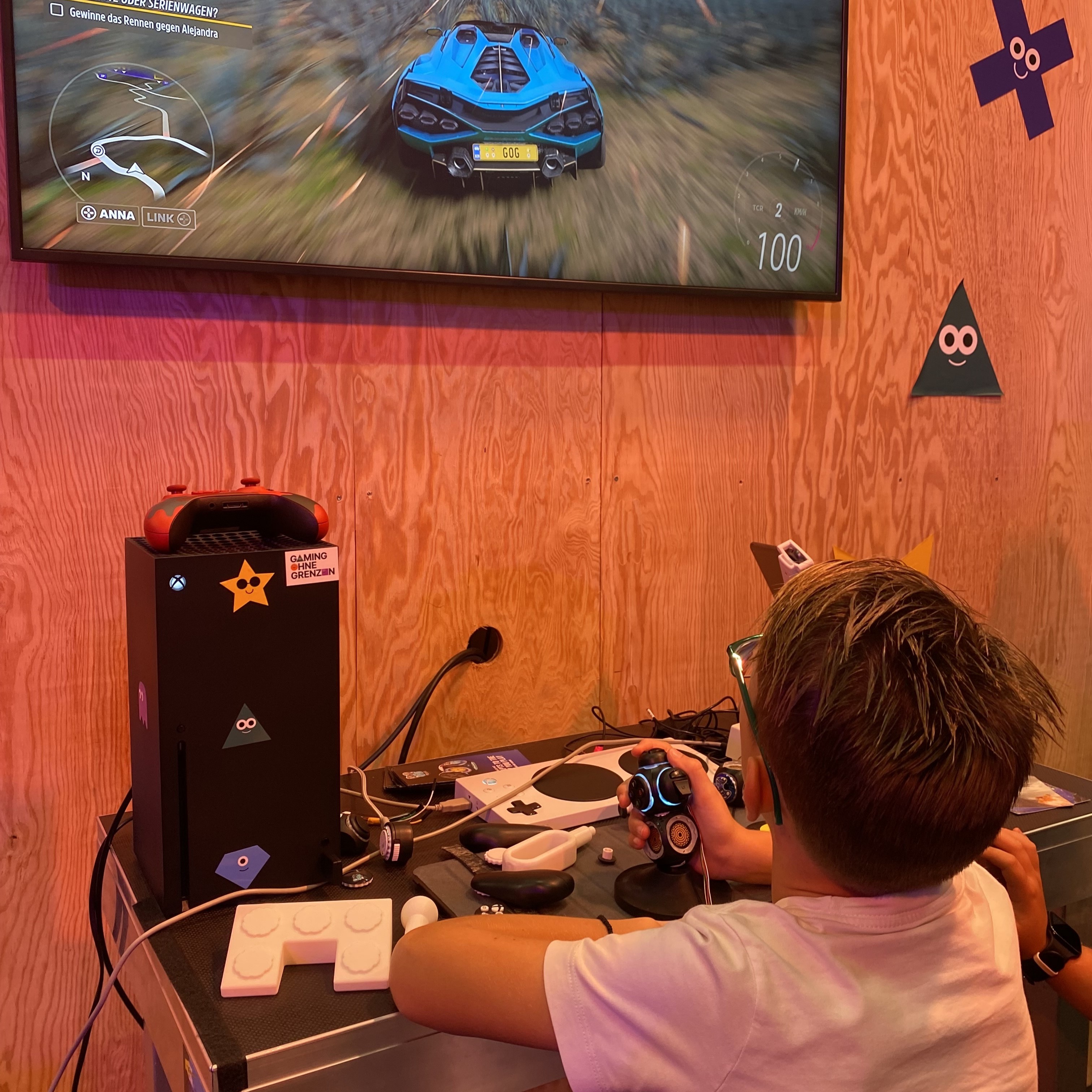 Ein Junge spielt Forza Horizon 5 mit einem Proteus Controller. Der Controller ist ein schwarzer Joystick mit Knöpfen. Er schaut auf einem Bildschirm vor ihn auf der Wand.