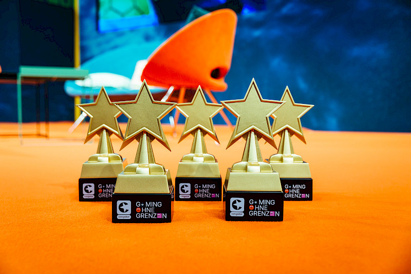 Die 5 Trophäen aus dem letzten Jahr stehen auf einem Teppich. Die Form der Trophäen sind Sterne auf einem Podest. Unten stehen die Logos von congstar und Gaming ohne Grenzen. Im Hintergrund stehen 2 Stühle und ein Tisch.
