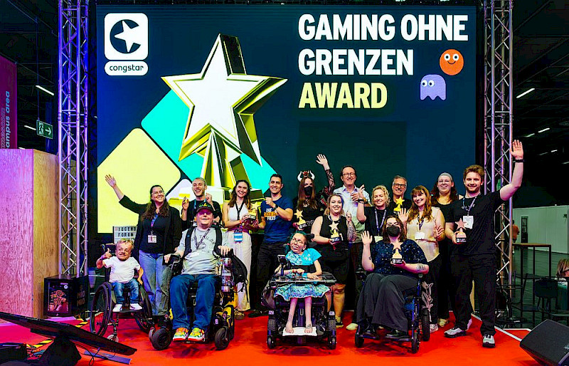 Ein Foto von allen Laudator*innen und Preisträger*innen des Gaming ohne Grenzen Awards 2025. Sie sind auf einer Bühne, lachen und manche winken in die Kamera. Im Hintergrund ist ein Bild projiziert. Auf dem Bild ist die Trophäe und das Logo von congstar.