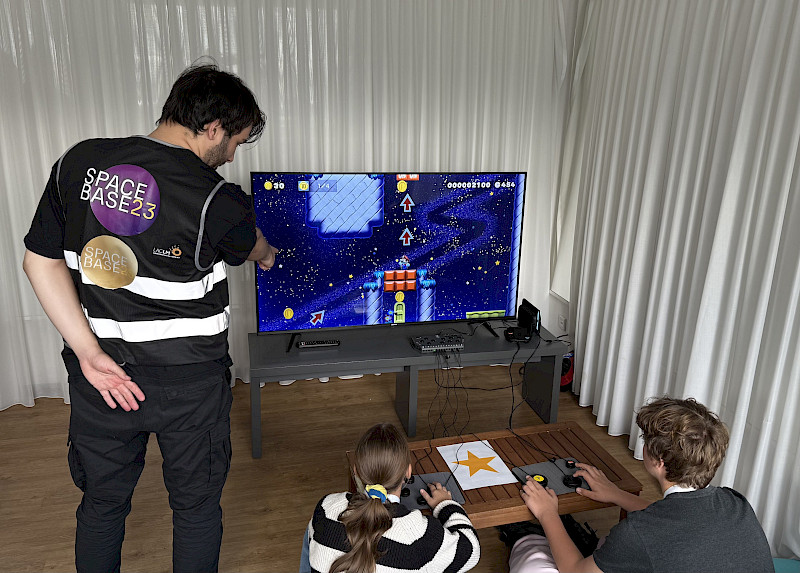 Zwei Jugendliche Spielen ein digitales Spiel. Ein Jugendlicher aus unserem Team steht da neben und zeigt auf etwas. Er trägt eine Weste mit der Aufschrift "SpaceBase 25".