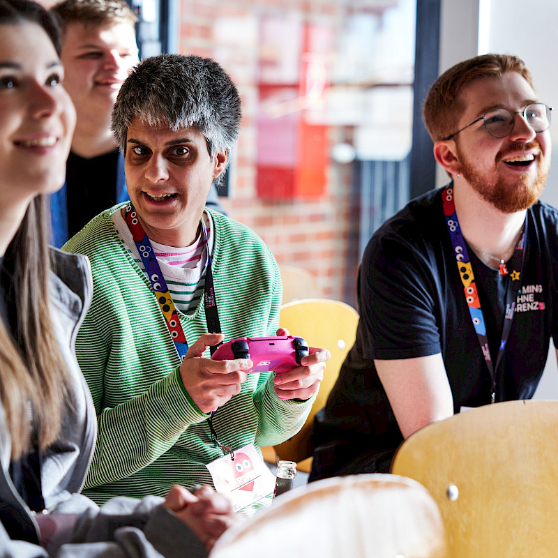 Mehrere Menschen sitzen zusammen in einem Raum. Eine Person in der Mitte hält einen Controller in der Hand und spielt ein Spiel. Die anderen schauen zu und lächeln. Alle tragen Namensschilder um den Hals. Die Stimmung wirkt freundlich und entspannt.