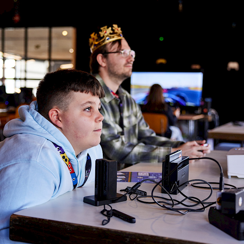 Zwei Menschen sitzen an einem Tisch und spielen ein Videospiel. Sie halten Controller in der Hand und schauen auf einen Bildschirm. Auf dem Tisch liegen Geräte und Kabel. Eine Person trägt eine Krone auf dem Kopf. Die Stimmung wirkt konzentriert und ruhig