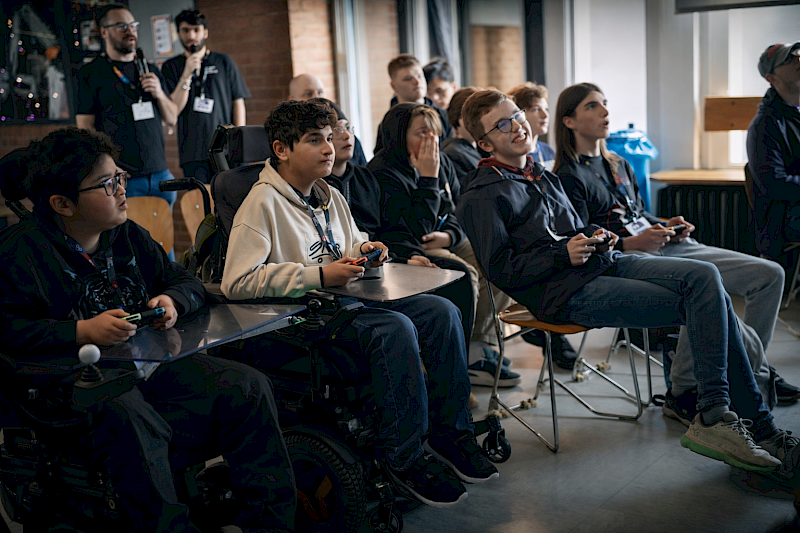 Mehrere Jugendliche sitzen in einem hellen Raum in einer Reihe und spielen gemeinsam Videospiele mit Controllern. Zwei von ihnen nutzen Rollstühle mit befestigten Ablagen für die Controller. Die Gruppe schaut konzentriert nach vorne auf einen Bildschirm a