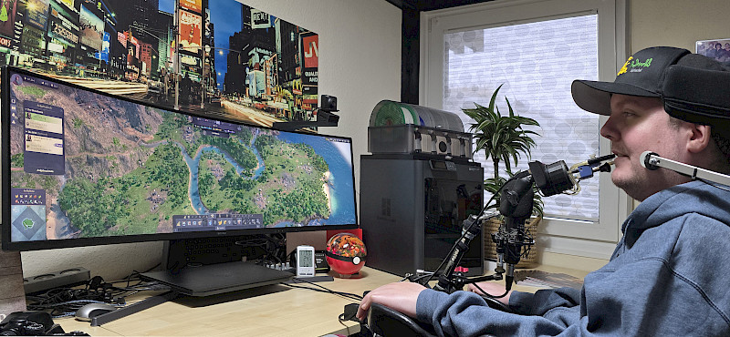 Eine Person sitzt an einem Schreibtisch vor zwei Monitoren und spielt ein Aufbaustrategie-Spiel. Sie trägt eine Kappe. Der Quadstcik ist an einer Halterung vor dem Mund befestigt. Neben den Monitoren stehen ein PC, eine Zimmerpflanze und verschiedene Gege