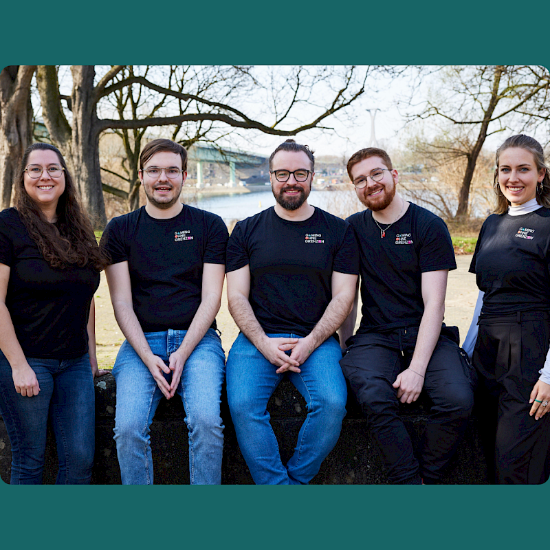 Fünf Menschen stehen und sitzen zusammen draußen. Drei sitzen auf einer niedrigen Mauer, zwei stehen daneben.  Alle tragen schwarze T-Shirts mit einem Logo. Sie schauen in die Kamera und lächeln. Im Hintergrund sieht man einen Fluss, eine Brücke und Bäume