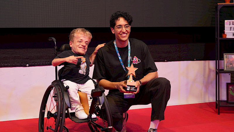 Zwei Männer präsentieren bei der „Gaming ohne Grenzen Award 2026“-Verleihung eine goldene Stern-Trophäe. Einer der beiden sitzt im Rollstuhl, der andere kniet neben ihm. Im Hintergrund ist die Bühnenwand der Veranstaltung zu sehen.